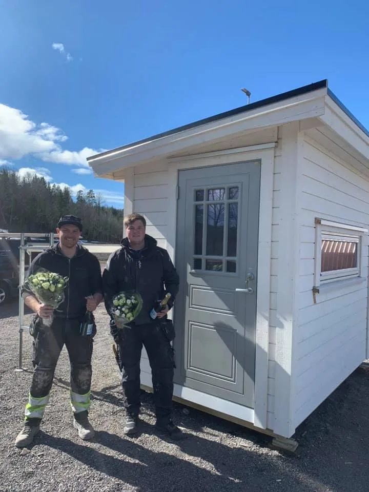 To nyutdannede tømrere med blomster foran svenneprøvestykket sitt, en hvit bod
